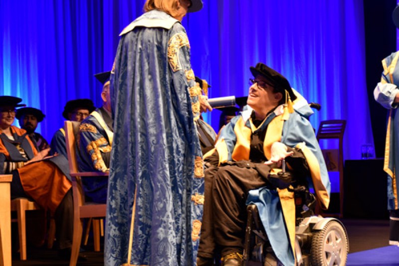 James Rose in his wheelchair, wearing ceremonial robes, being handed a certificate from a lady also in ceremonial robes